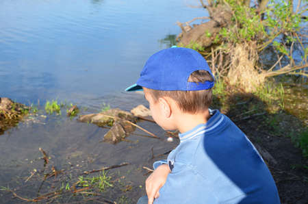 A boy stands by the forest lake and looks at the calm water. Nature, contemplation, inspiration, outdoor. Walk outdoors. loneliness. young cute boys fishing on a lake in a sunny summer or spring day.の写真素材