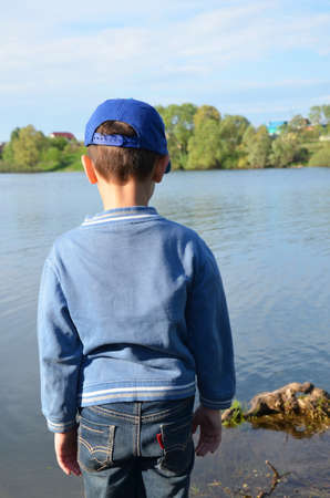 A boy stands by the forest lake and looks at the calm water. Nature, contemplation, inspiration, outdoor. Walk outdoors. loneliness. young cute boys fishing on a lake in a sunny summer or spring day.の写真素材