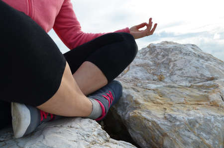 young woman meditation in a yoga pose at the beach. girl in lotus position on an empty stone seashore. takes yoga, sports, meditation. healthy lifestyle. close up sneakers sportswear.の写真素材