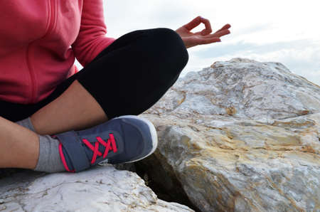 young woman meditation in a yoga pose at the beach. girl in lotus position on an empty stone seashore. takes yoga, sports, meditation. healthy lifestyle. close up sneakers sportswear.の写真素材