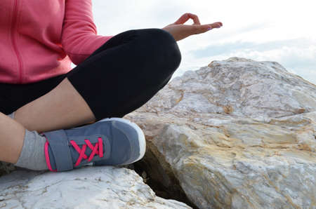 young woman meditation in a yoga pose at the beach. girl in lotus position on an empty stone seashore. takes yoga, sports, meditation. healthy lifestyle. close up sneakers sportswear.の写真素材