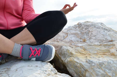 young woman meditation in a yoga pose at the beach. girl in lotus position on an empty stone seashore. takes yoga, sports, meditation. healthy lifestyle. close up sneakers sportswear.の写真素材