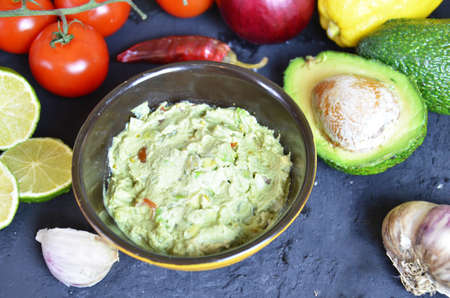 Bowl of freshly prepared guacamole. Ingredients on the background tomatoes, hot pepper, lime, avocado Guacamole nachos and guacamole ingredients on dark wooden background.の写真素材