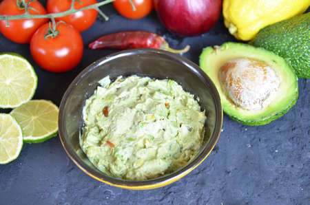 Bowl of freshly prepared guacamole. Ingredients on the background tomatoes, hot pepper, lime, avocado Guacamole nachos and guacamole ingredients on dark wooden background.の写真素材