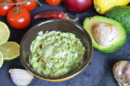 Bowl of freshly prepared guacamole. Ingredients on the background tomatoes, hot pepper, lime, avocado Guacamole nachos and guacamole ingredients on dark wooden background.の写真素材