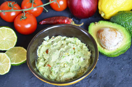 Bowl of freshly prepared guacamole. Ingredients on the background tomatoes, hot pepper, lime, avocado Guacamole nachos and guacamole ingredients on dark wooden background.の写真素材