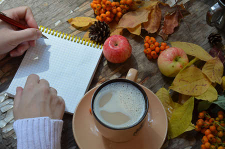 Workspace with golden maple leaves, notebook, coffee cup. Stylish office desk. Autumn or Winter concept. Flat lay, top view. A female hand in a white knitted suite writes in a red handmade notebook, dry autumn leaves, apples and berries next to a cup of cappuccino coffee.の写真素材