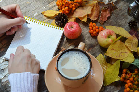 Workspace with golden maple leaves, notebook, coffee cup. Stylish office desk. Autumn or Winter concept. Flat lay, top view. A female hand in a white knitted suite writes in a red handmade notebook, dry autumn leaves, apples and berries next to a cup of cappuccino coffee.の写真素材