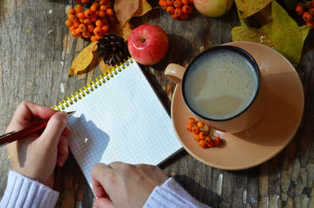 Workspace with golden maple leaves, notebook, coffee cup. Stylish office desk. Autumn or Winter concept. Flat lay, top view. A female hand in a white knitted suite writes in a red handmade notebook, dry autumn leaves, apples and berries next to a cup of cappuccino coffee.の写真素材