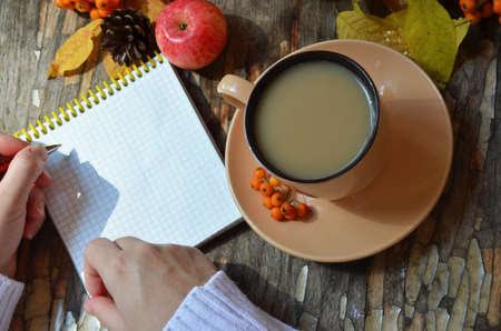 Workspace with golden maple leaves, notebook, coffee cup. Stylish office desk. Autumn or Winter concept. Flat lay, top view. A female hand in a white knitted suite writes in a red handmade notebook, dry autumn leaves, apples and berries next to a cup of cappuccino coffee.の写真素材