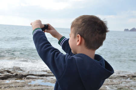 boy schoolboy or teenager in a dark blue tracksuit walks along the sea, takes a selfie or shoots the sea on an action camera, on the sea waves, stone coast, wind.の写真素材