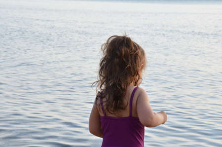 a little cute, very cute girl stands near the evening sea and looks at the water. Pensive, serious. in a purple swimsuit. on the beach near sea, summer, vacation, travel concept. Baby girlの写真素材