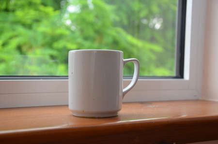 A white cup of tea or coffee stands on a wooden window sill against the background of a summer...