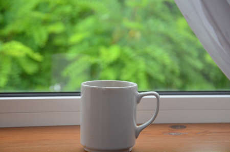 A white cup of tea or coffee stands on a wooden window sill against the background of a summer garden.の写真素材