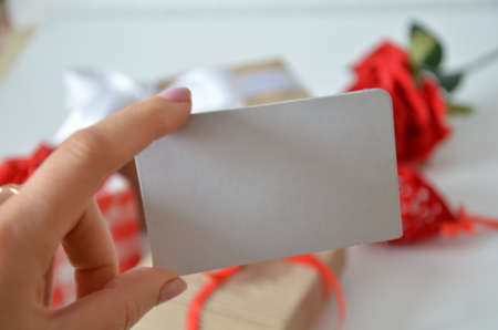 Girl holds a blank card without an inscription on the background of gifts with bows, ribbons and red rose on a white background. concept of discounts, gift, mothers day, womens day, valentines dayの写真素材