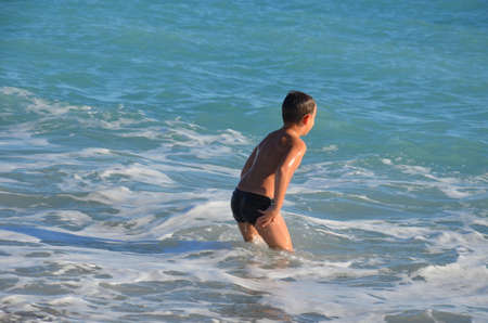 A small, happy boy stands in the water and splashes. A cheerful child jumps on the sea shore, the beach. The child enjoys the sea and splashes in the water with cottage cheese. Rear viewの写真素材