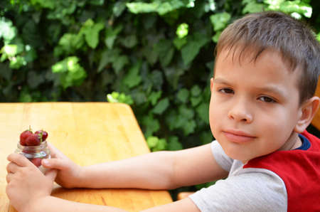 cute little boys hold in their hand a jar of yogurt or pudding with their strawberries and cherries, in nature, on a background of green foliage Breakfast, healthy food, happy childhood. sitting eating greek yogurt.の写真素材
