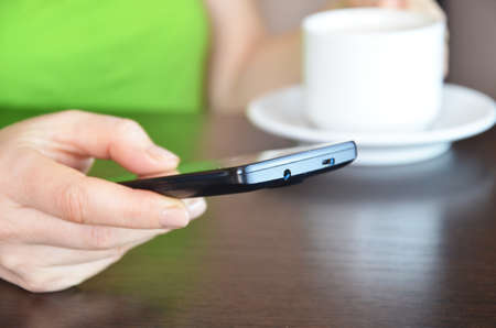 girl holds a phone in her hands and a cup of coffee. morning. against the window. morning starts on the phone. chatting news. Close up of hands woman using her cell phone in homeの写真素材