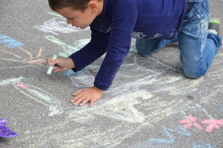 Little child boy smiling siting on asphalt sidewalk,drawing house, sun painted by colored chalk.Children's picture,creativity on gray dackground road. Kid looking camera, top view,copy space.の写真素材