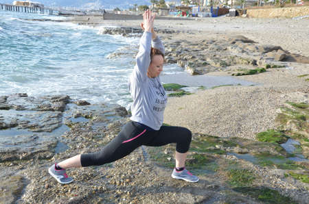 Beautiful blond girl clothing in at the seaside on the rock doing yoga poses, stretching or sports gymnastics and meditating in yoga pose.の写真素材