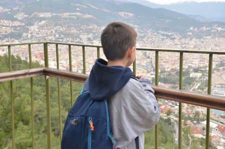 Little boy, looking at the amazing view on the Mediterranian sea gulf with touristic ships and aincient castle and a city from the mountain with a backpack on the backの写真素材