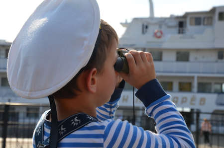Little boy in sailors uniform with binocular in the boat A boy in a jacket matrsa cap looks at the sea through binoculars at the sea near the shipsの写真素材