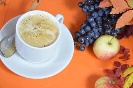 White cup of coffee with cappuccino cream on an orange background with autumn leaves, apples and red berries. Fall season, leisure time and coffee break concept.の写真素材