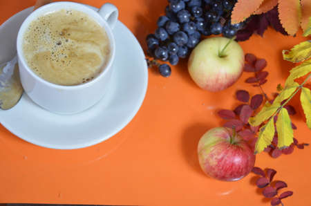 White cup of coffee with cappuccino cream on an orange background with autumn leaves, apples and red berries. Fall season, leisure time and coffee break concept.の写真素材