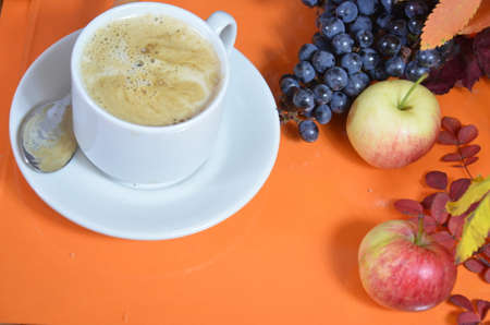 White cup of coffee with cappuccino cream on an orange background with autumn leaves, apples and red berries. Fall season, leisure time and coffee break concept.の写真素材