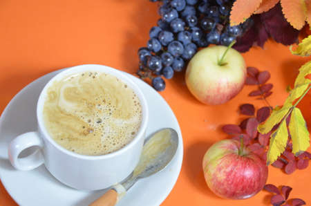 White cup of coffee with cappuccino cream on an orange background with autumn leaves, apples and red berries. Fall season, leisure time and coffee break concept.の写真素材