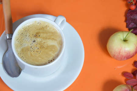 White cup of coffee with cappuccino cream on an orange background with autumn leaves, apples and red berries. Fall season, leisure time and coffee break concept. Autumn leaves and hot steaming coffeeの写真素材