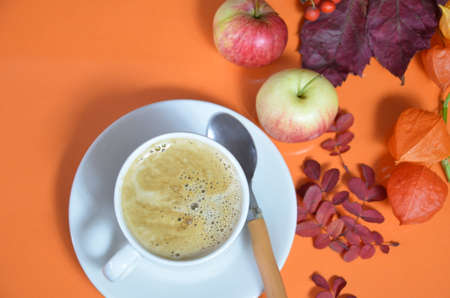 White cup of coffee with cappuccino cream on an orange background with autumn leaves, apples and red berries. Fall season, leisure time and coffee break concept. Autumn leaves and hot steaming coffeeの写真素材