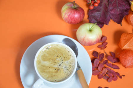 White cup of coffee with cappuccino cream on an orange background with autumn leaves, apples and red berries. Fall season, leisure time and coffee break concept. Autumn leaves and hot steaming coffeeの写真素材