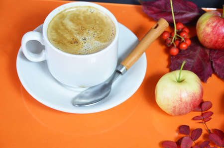 White cup of coffee with cappuccino cream on an orange background with autumn leaves, apples and red berries. Fall season, leisure time and coffee break concept. Autumn leaves and hot steaming coffeeの写真素材