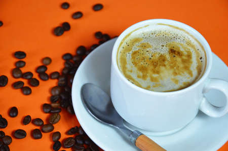 warm cup of coffee cappuccino with cream in a white cup on an orange and black background with coffee beans with a teaspoonの写真素材