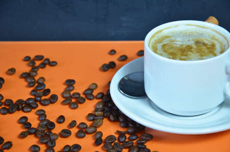 warm cup of coffee cappuccino with cream in a white cup on an orange and black background with coffee beans with a teaspoonの写真素材