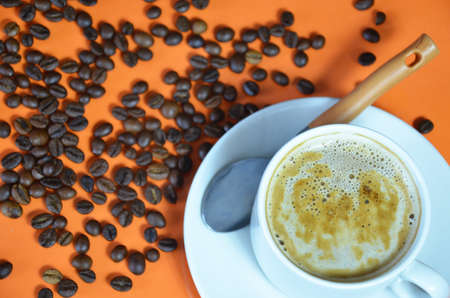 warm cup of coffee cappuccino with cream in a white cup on an orange and black background with coffee beans with a teaspoonの写真素材