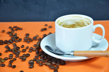 warm cup of coffee cappuccino with cream in a white cup on an orange and black background with coffee beans with a teaspoonの写真素材