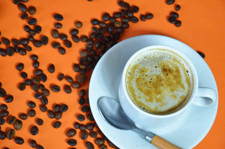 warm cup of coffee cappuccino with cream in a white cup on an orange and black background with coffee beans with a teaspoonの写真素材