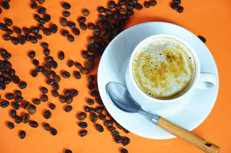 warm cup of coffee cappuccino with cream in a white cup on an orange and black background with coffee beans with a teaspoonの写真素材