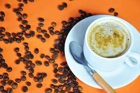 warm cup of coffee cappuccino with cream in a white cup on an orange and black background with coffee beans with a teaspoonの写真素材