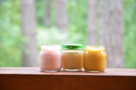 Three jars of baby food nutrition puree mash on the background of foliage. Pink light yellow orange colorの写真素材