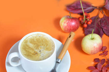 White cup of coffee with cappuccino cream on an orange background with autumn leaves, apples and red berries. Fall season, leisure time and coffee break concept. Autumn leaves and hot steaming coffeeの写真素材