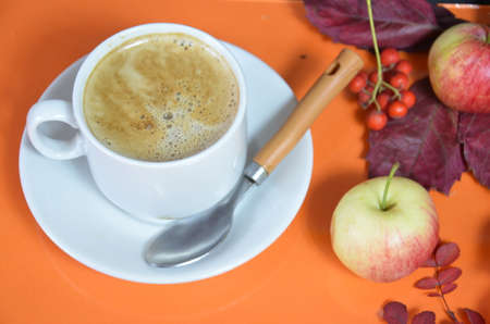 White cup of coffee with cappuccino cream on an orange background with autumn leaves, apples and red berries. Fall season, leisure time and coffee break concept. Autumn leaves and hot steaming coffeeの写真素材