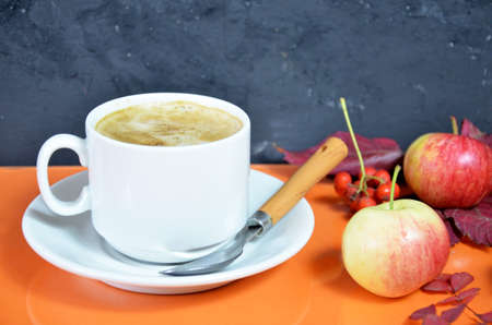 White cup of coffee with cappuccino cream on an orange background with autumn leaves, apples and red berries. Fall season, leisure time and coffee break concept. Autumn leaves and hot steaming coffee.の写真素材