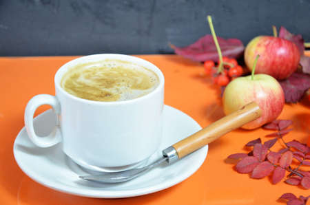 White cup of coffee with cappuccino cream on an orange background with autumn leaves, apples and red berries. Fall season, leisure time and coffee break concept. Autumn leaves and hot steaming coffeeの写真素材