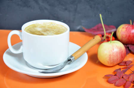 White cup of coffee with cappuccino cream on an orange background with autumn leaves, apples and red berries. Fall season, leisure time and coffee break concept. Autumn leaves and hot steaming coffeeの写真素材