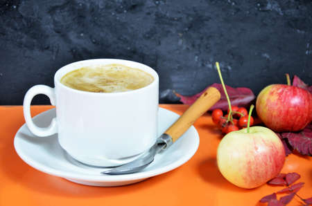 White cup of coffee with cappuccino cream on an orange background with autumn leaves, apples and red berries. Fall season, leisure time and coffee break concept. Autumn leaves and hot steaming coffeeの写真素材