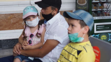 Father with daughter in his arms, son sits wearing masks at station or at the airport, waiting for the train. Family in face mask. Virus. Coronavirus and flu pandemic. Safe travel with child and babyの写真素材