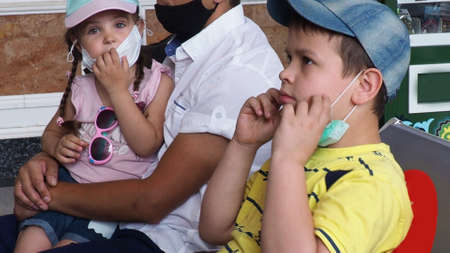 Father with daughter in his arms, son sits wearing masks at station or at the airport, waiting for the train. Family in face mask. Virus. Coronavirus and flu pandemic. Safe travel with child and babyの写真素材